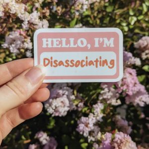 Hånd som holder klistremerke med teksten: Hello, I'm Disassociating. Blomster i bakgrunnen.
